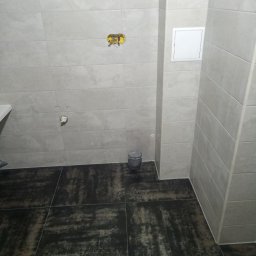 Wykończona łazienka w Bolesławcu z beżowymi płytkami ściennymi i ciemną podłogą. Widoczne przyłącza hydrauliczne i elektryczne, gotowe do montażu armatury.