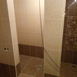 Wykończona kabina prysznicowa z beżowo-brązowymi płytkami, widoczny odpływ liniowy i fragment instalacji elektrycznej. Ukończone prace glazurnicze w łazience.
