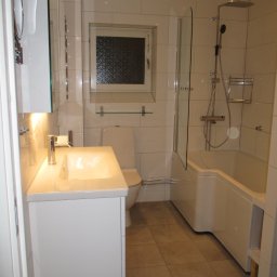 Biała, nowoczesna łazienka z wanną, umywalką i toaletą. Jasne płytki na ścianach i podłodze. Minimalistyczny design z oświetleniem punktowym i lustrem.