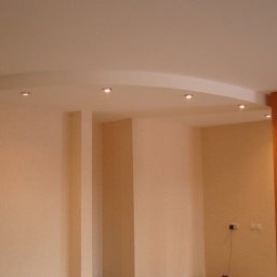 Remont Bud Expert - Jasne wnętrze z podwieszanym sufitem o łukowatym kształcie i wbudowanym oświetleniem punktowym. Ściany w odcieniach beżu i brązu, widoczne gniazdka elektryczne.