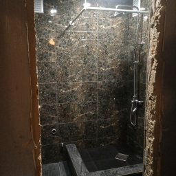 Remont Bud Expert - Wykończona kabina prysznicowa z ciemnymi, marmurkowymi płytkami, widoczna przez surowe, nieotynkowane ościeżnice. Chromowana armatura i kratka odpływowa.