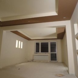 Remont Bud Expert - Wykończone wnętrze pokoju z dekoracyjnym sufitem podwieszanym w odcieniach bieli i brązu. Widoczne okno z grzejnikiem i wnęki w ścianie.