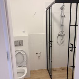 Remont Bud Expert - Nowoczesna łazienka po remoncie: kabina prysznicowa z czarnymi ramami, toaleta z podtynkową spłuczką, jasne płytki na ścianach i podłodze imitującej drewno.