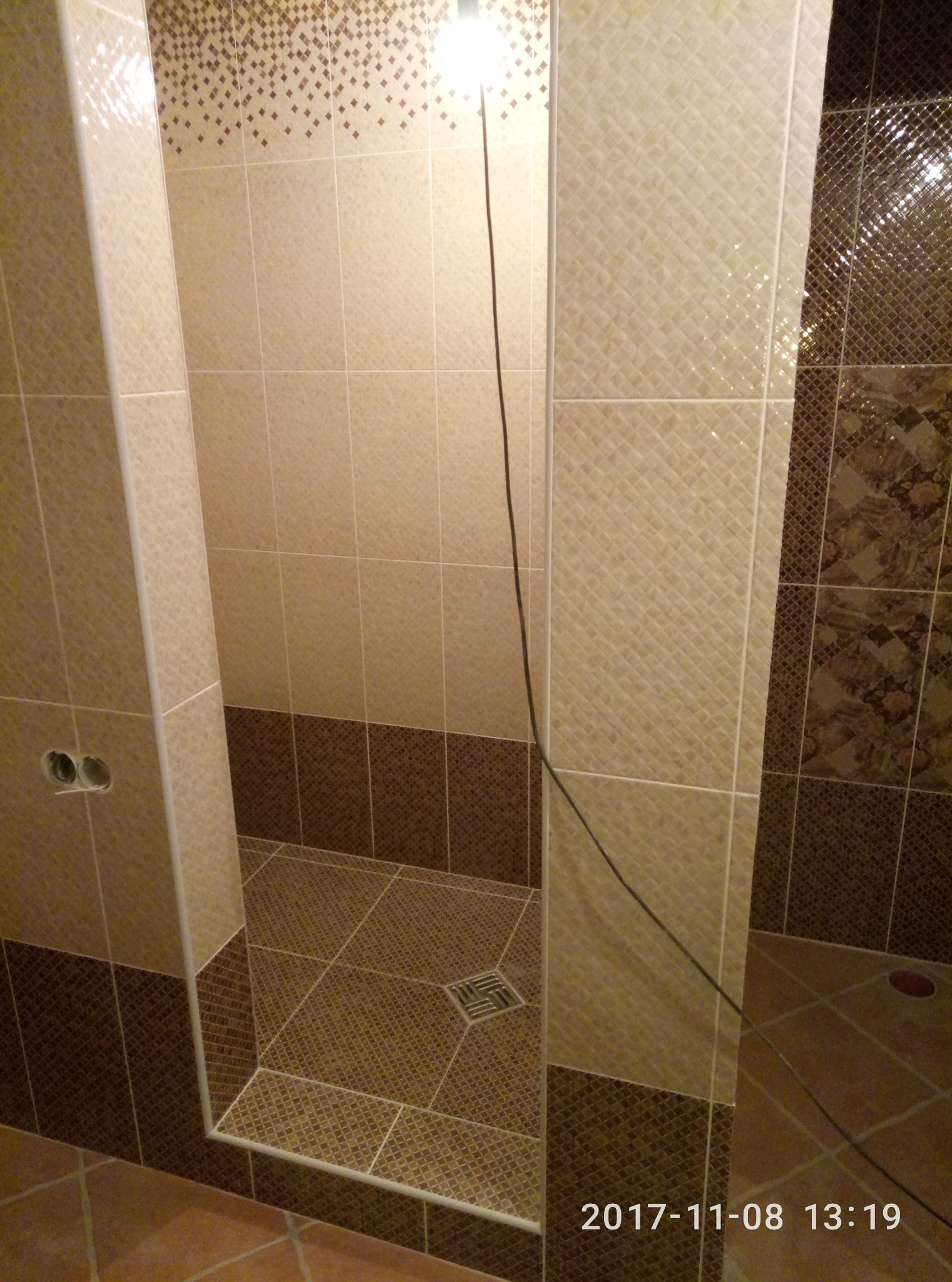 Wykończona kabina prysznicowa z beżowo-brązowymi płytkami, widoczny odpływ liniowy i fragment instalacji elektrycznej. Ukończone prace glazurnicze w łazience.