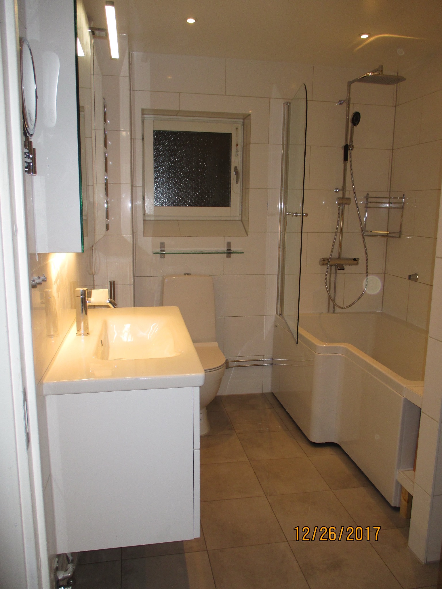 Biała, nowoczesna łazienka z wanną, umywalką i toaletą. Jasne płytki na ścianach i podłodze. Minimalistyczny design z oświetleniem punktowym i lustrem.