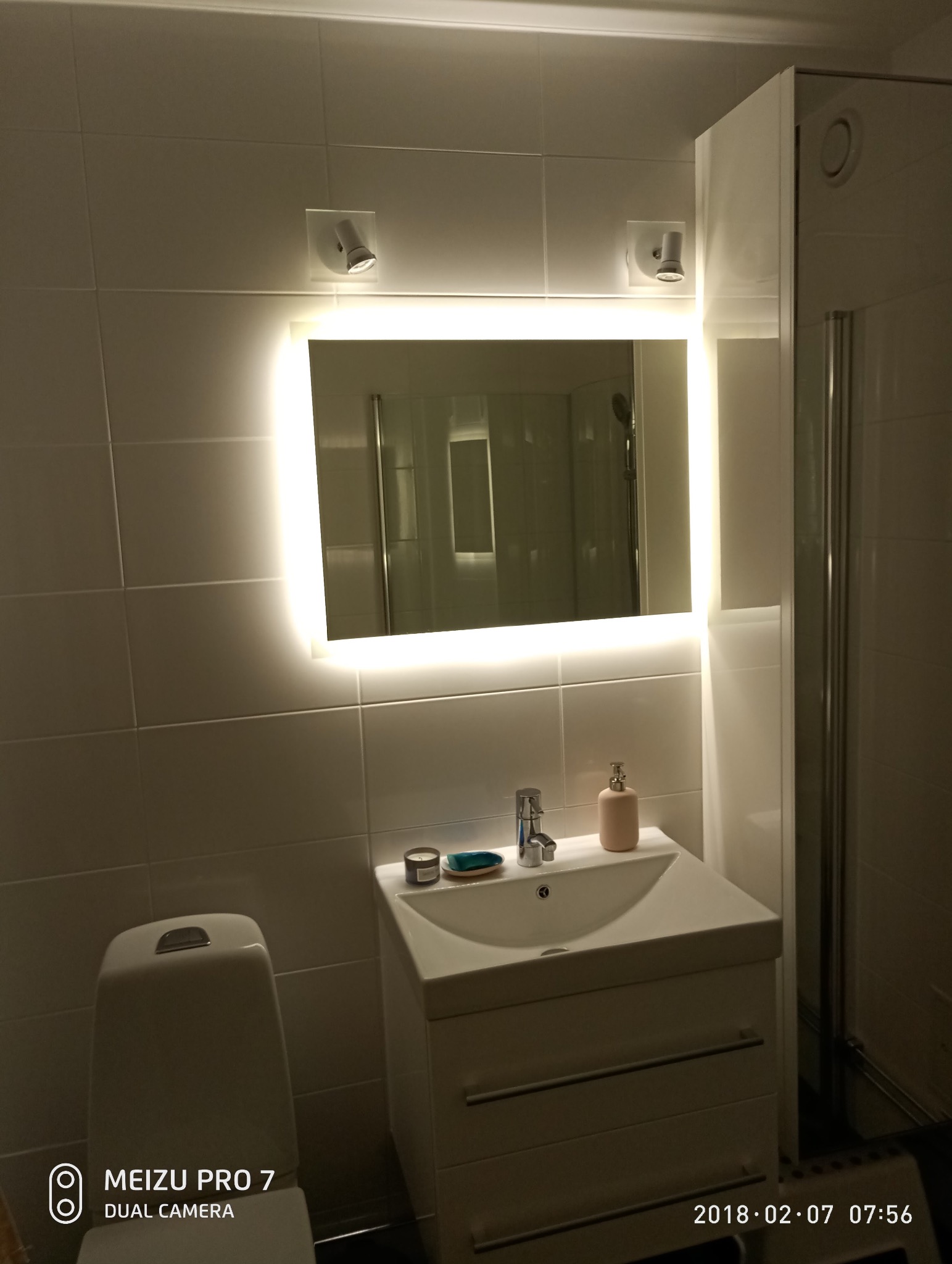 Biała łazienka z podświetlanym lustrem, umywalką z szafką i toaletą. Dwa reflektory nad lustrem. Widoczna kabina prysznicowa. Minimalistyczny design.