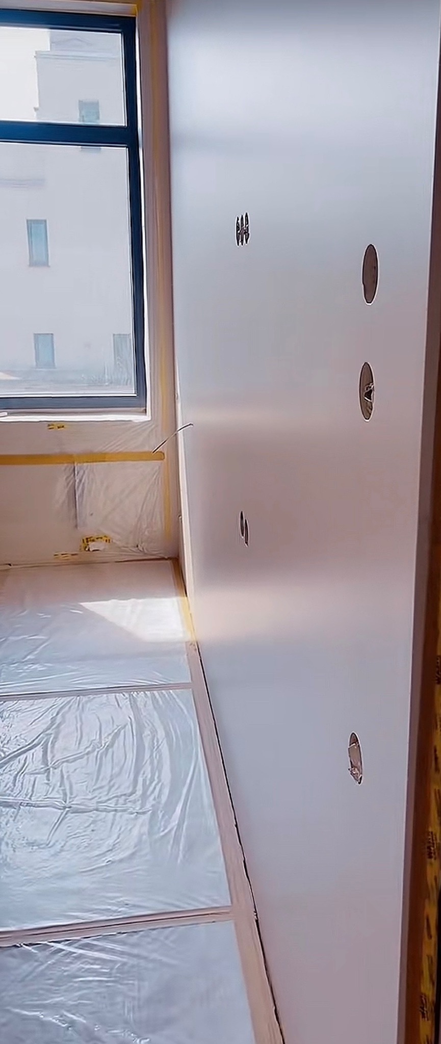 Biała ściana z otworami na gniazdka elektryczne w trakcie prac wykończeniowych. Podłoga i okno zabezpieczone folią malarską.