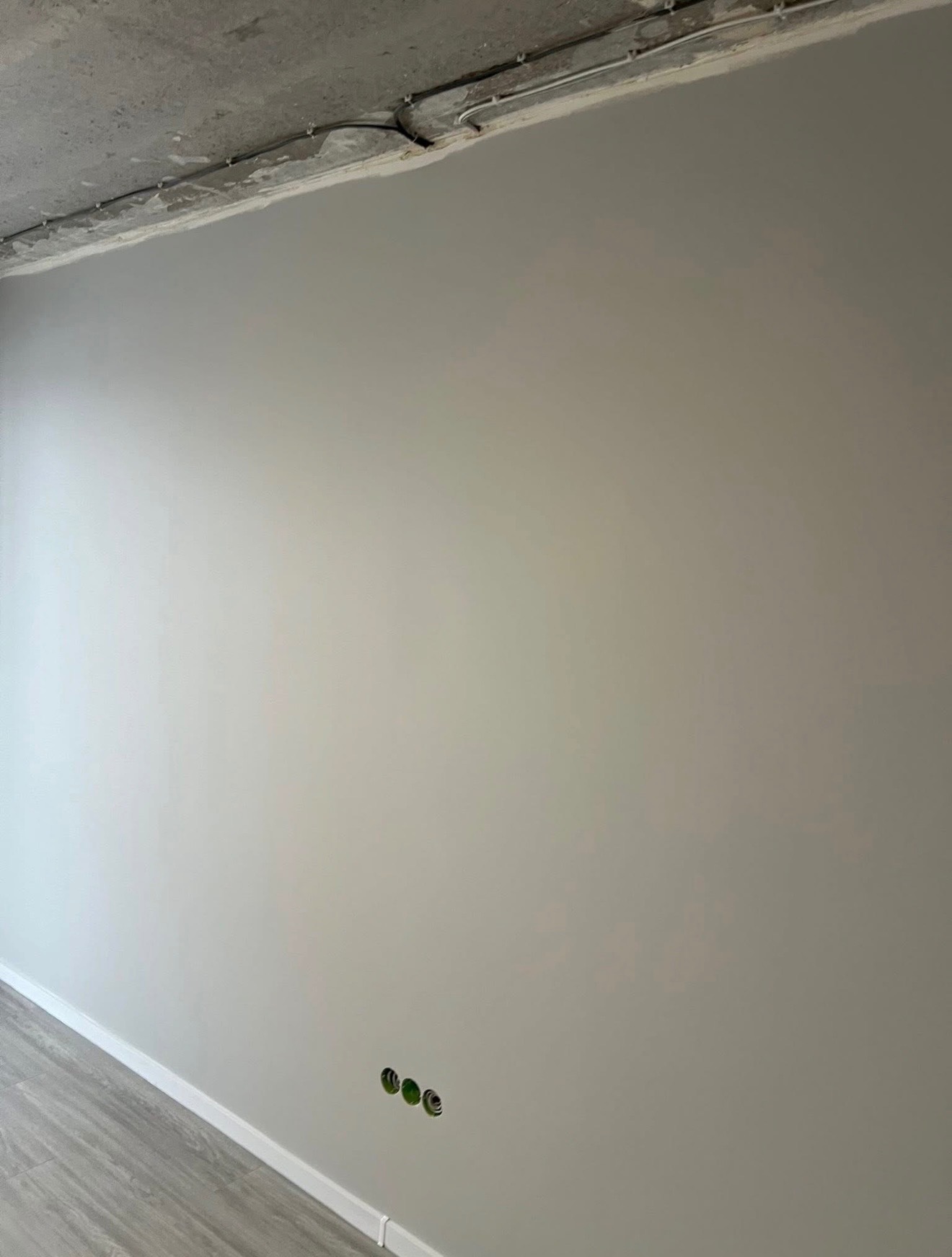 Gładka, pomalowana na szaro ściana z trzema gniazdkami elektrycznymi w dolnym rogu, widoczne szare panele podłogowe i surowy betonowy sufit z instalacją.