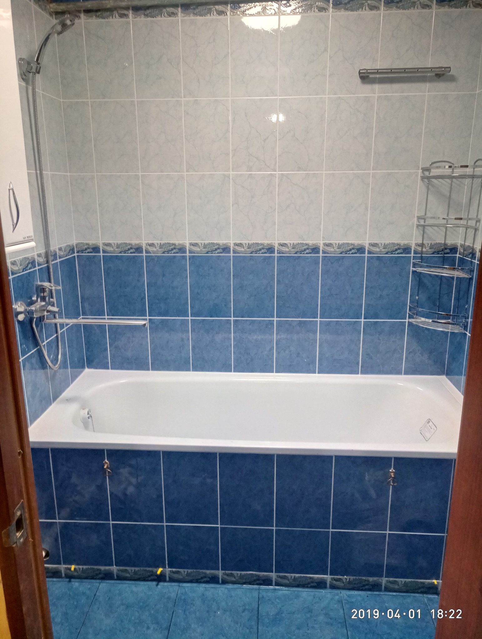 Wyremontowana łazienka z wanną obłożoną niebieskimi płytkami, nad nią jasne płytki. Widoczna bateria, słuchawka prysznicowa i półka na kosmetyki. Ujęcie z poziomu podłogi.