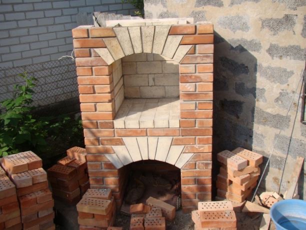 Murowany grill ogrodowy w trakcie budowy, z cegły klinkierowej i jasnych elementów dekoracyjnych. Widoczne cegły przygotowane do dalszej pracy.