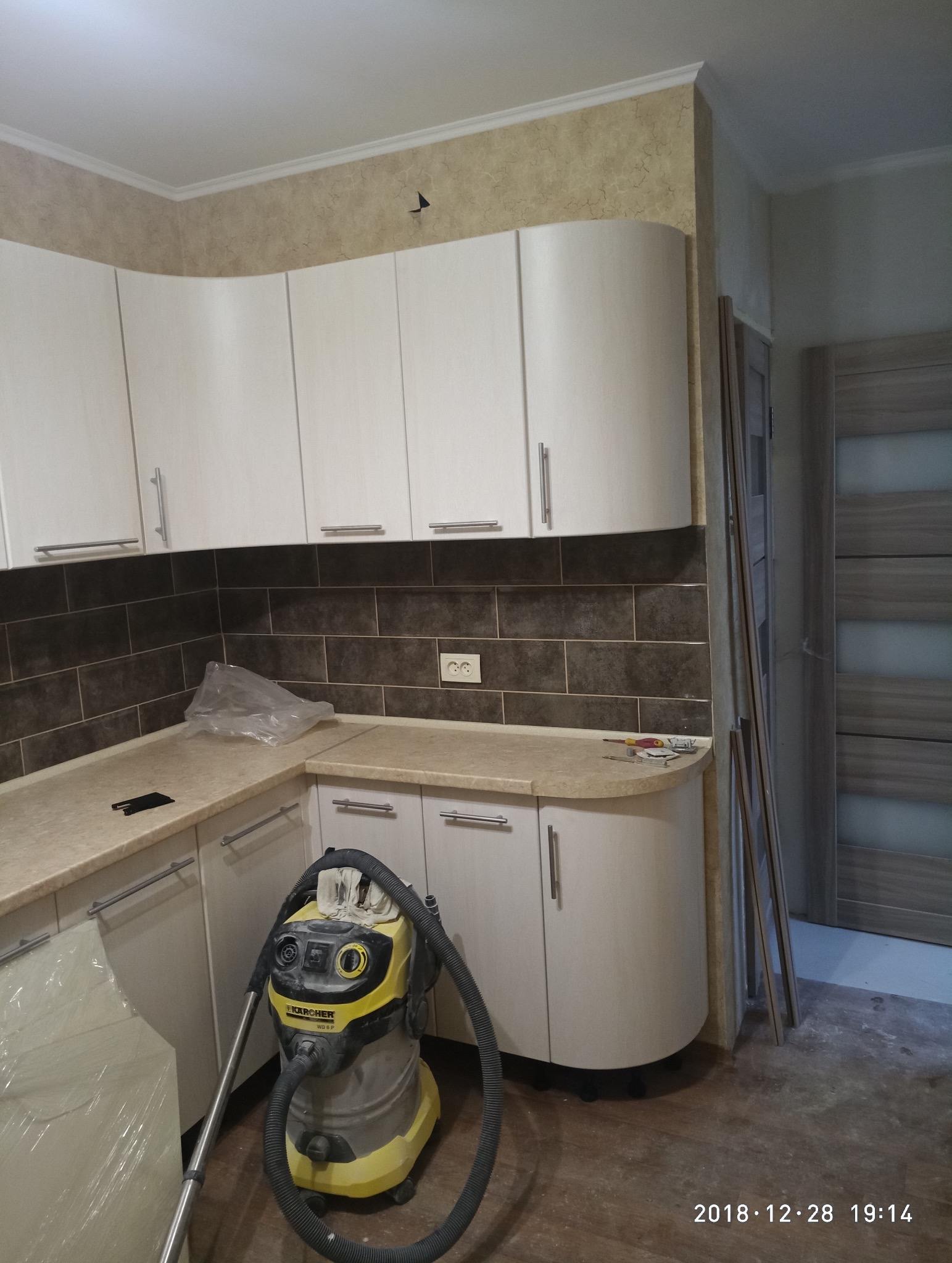 Jasna zabudowa kuchenna z brązowymi płytkami nad blatem. Na blacie leżą narzędzia, a na podłodze stoi odkurzacz przemysłowy. Widoczny fragment drzwi.