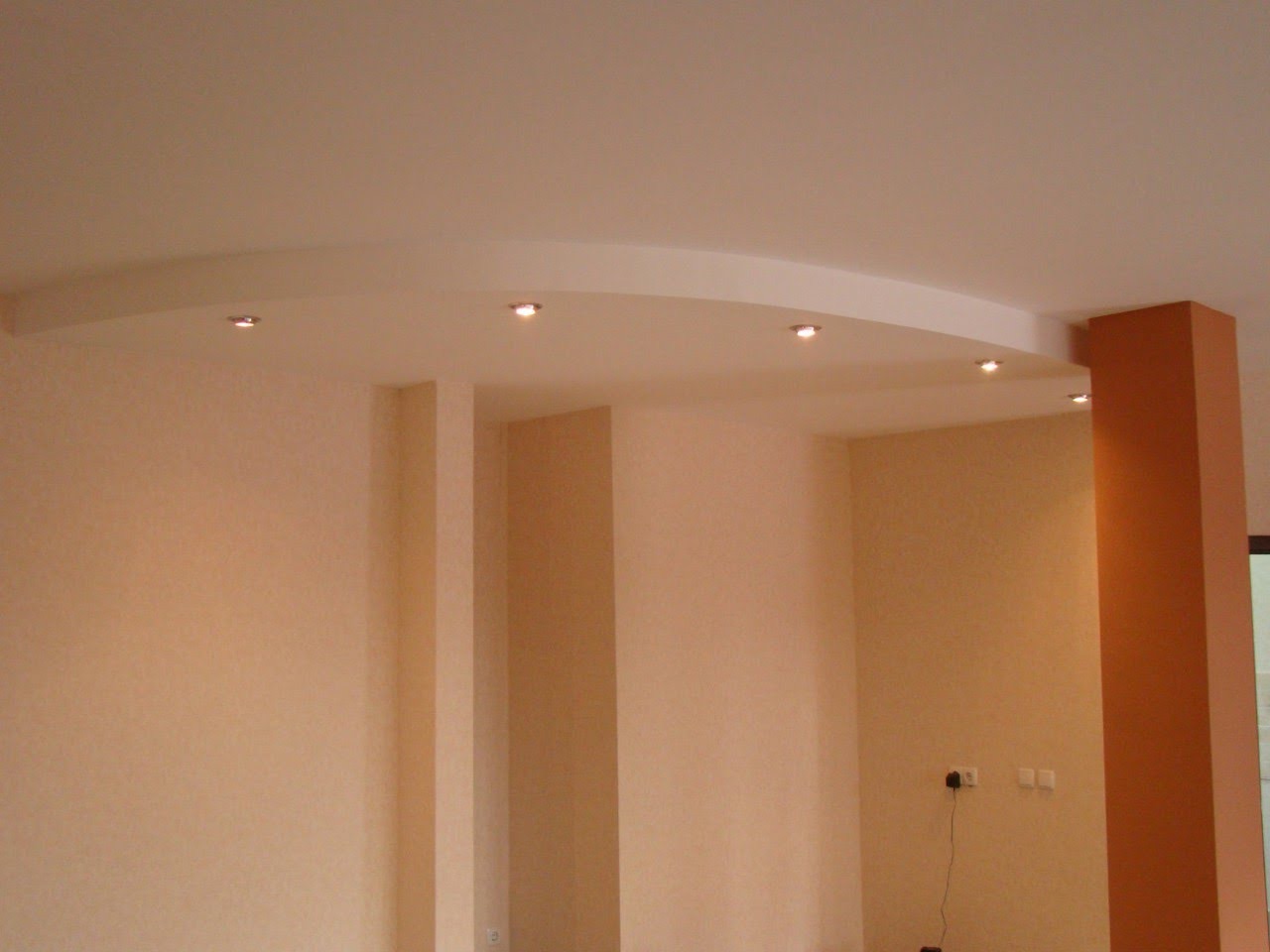 Jasne wnętrze z podwieszanym sufitem o łukowatym kształcie i wbudowanym oświetleniem punktowym. Ściany w odcieniach beżu i brązu, widoczne gniazdka elektryczne.