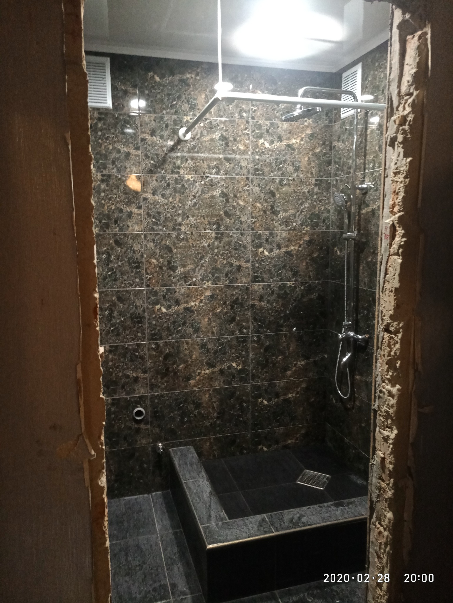 Wykończona kabina prysznicowa z ciemnymi, marmurkowymi płytkami, widoczna przez surowe, nieotynkowane ościeżnice. Chromowana armatura i kratka odpływowa.