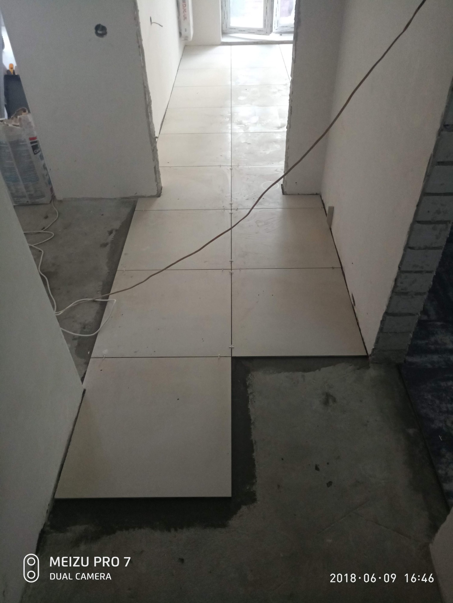 Układanie jasnych, kwadratowych płytek podłogowych w korytarzu. Widoczne krzyżyki dystansowe i fragmenty niezapełnionej przestrzeni pod płytkami. Ściany przygotowane do dalszych prac.