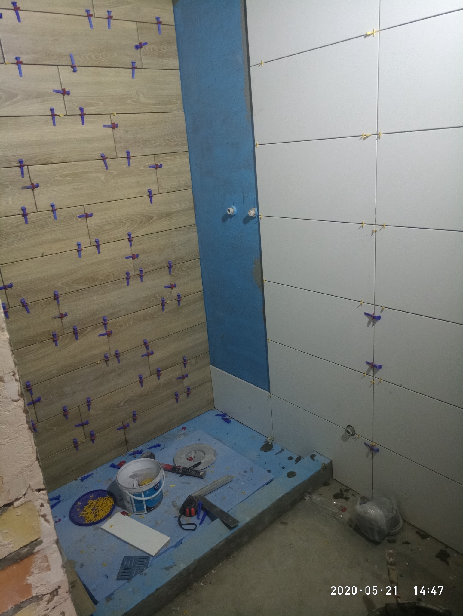 Układanie płytek w łazience: jasne płytki imitujące drewno i białe, poziome płytki. Widoczne narzędzia glazurnicze i niebieska hydroizolacja. Etap prac wykończeniowych.