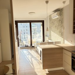 Volodymyr Vihor - Jasne, nowoczesne wnętrze z balkonem. Parkiet w odcieniach beżu, drewniane meble kuchenne i dekoracyjna tapeta z motywem kwiatowym. Widok na budynki zza okna.