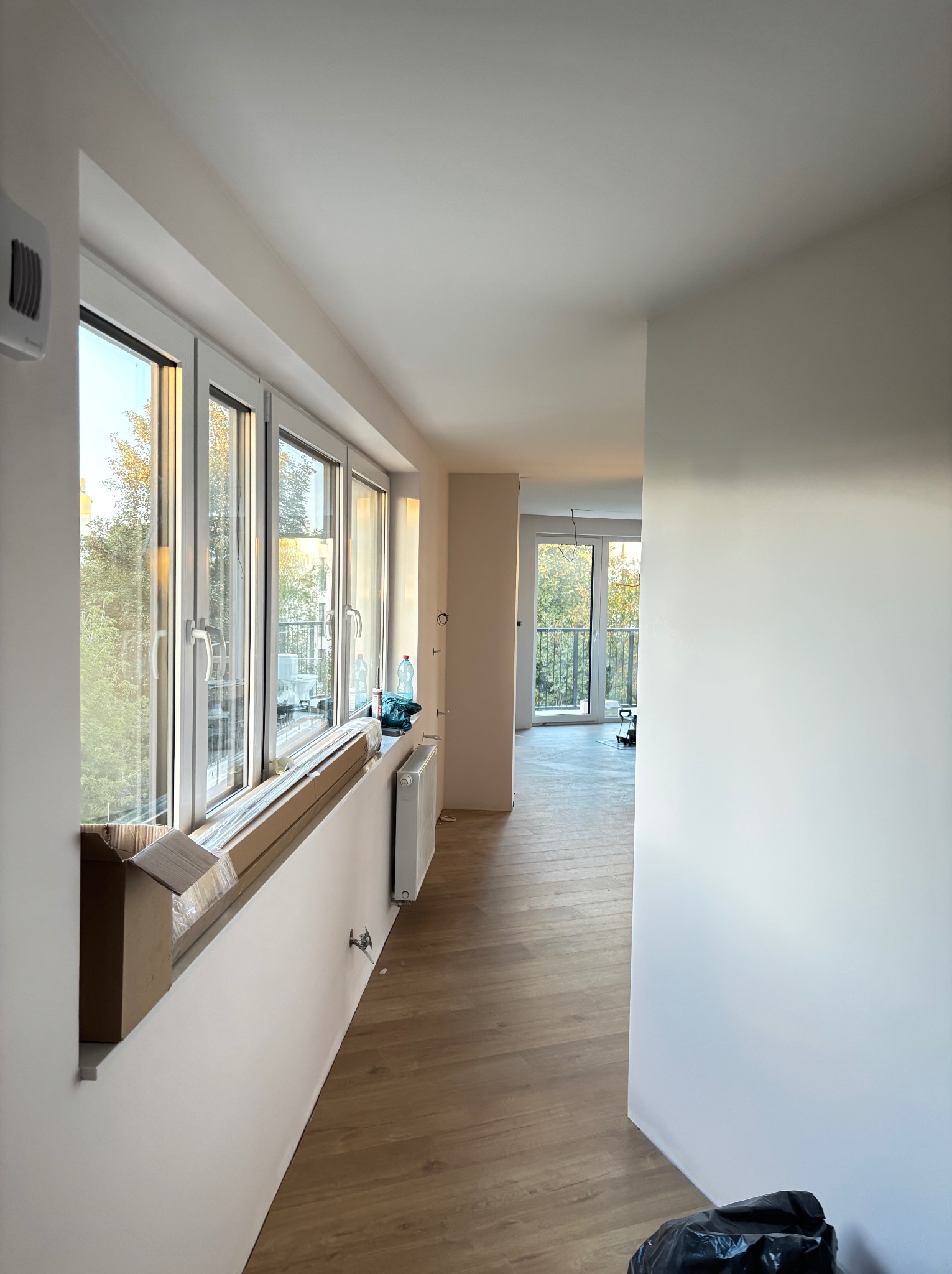 Jasne wnętrze w trakcie prac wykończeniowych: świeżo pomalowane ściany, ułożona podłoga z paneli, okna z widokiem na zieleń. Częściowo widoczne narzędzia i materiały.