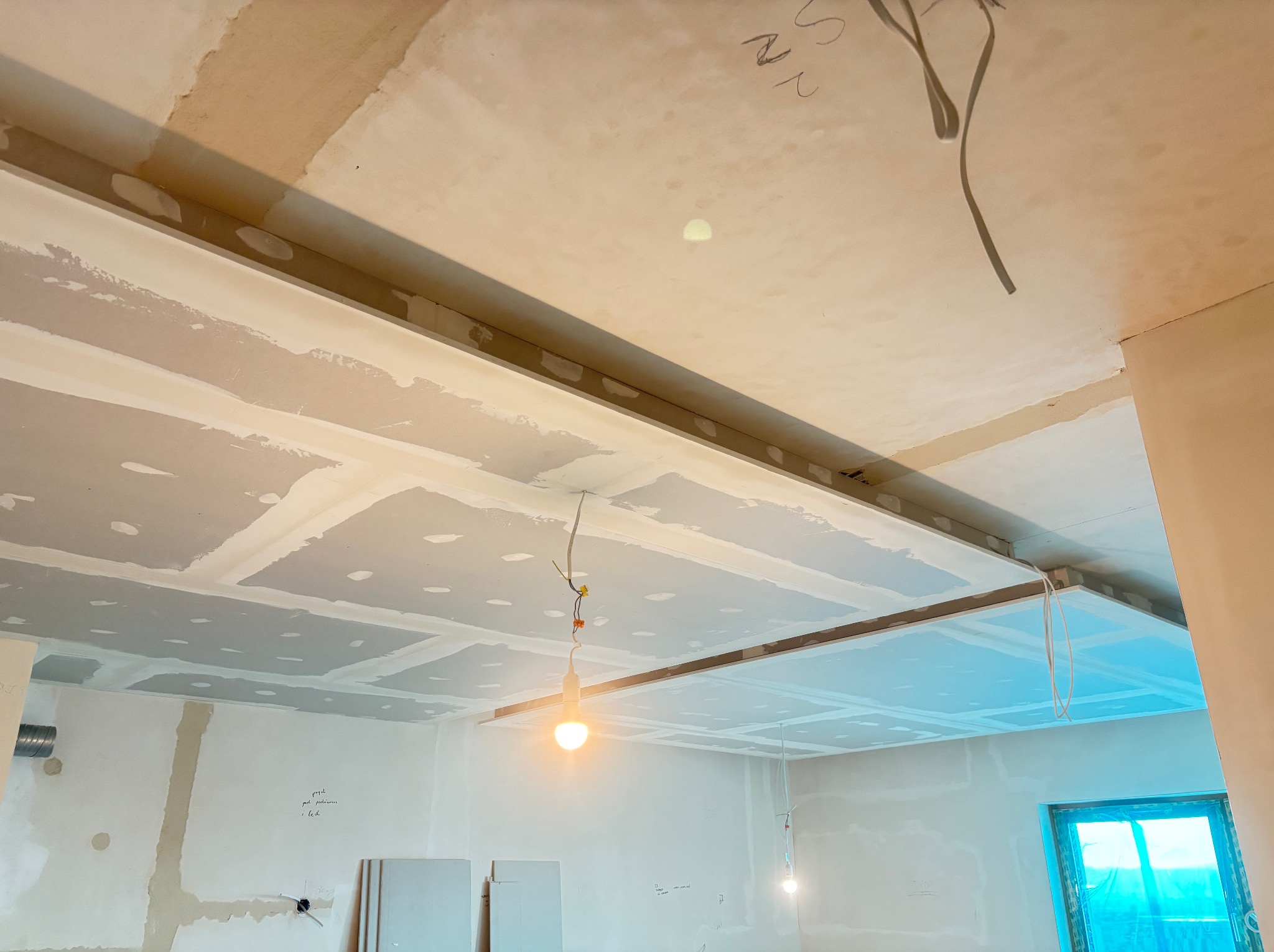 Sufit podwieszany z płyt karton-gips, widoczne łączenia i szpachlowanie. W tle ściany w trakcie przygotowania do malowania, okno z niebieską folią ochronną.