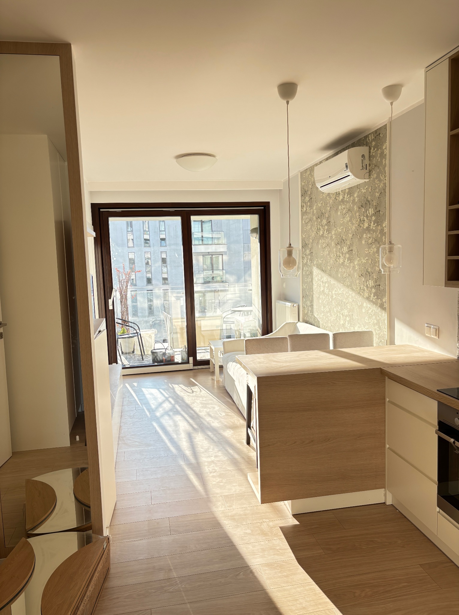 Jasne, nowoczesne wnętrze z balkonem. Parkiet w odcieniach beżu, drewniane meble kuchenne i dekoracyjna tapeta z motywem kwiatowym. Widok na budynki zza okna.