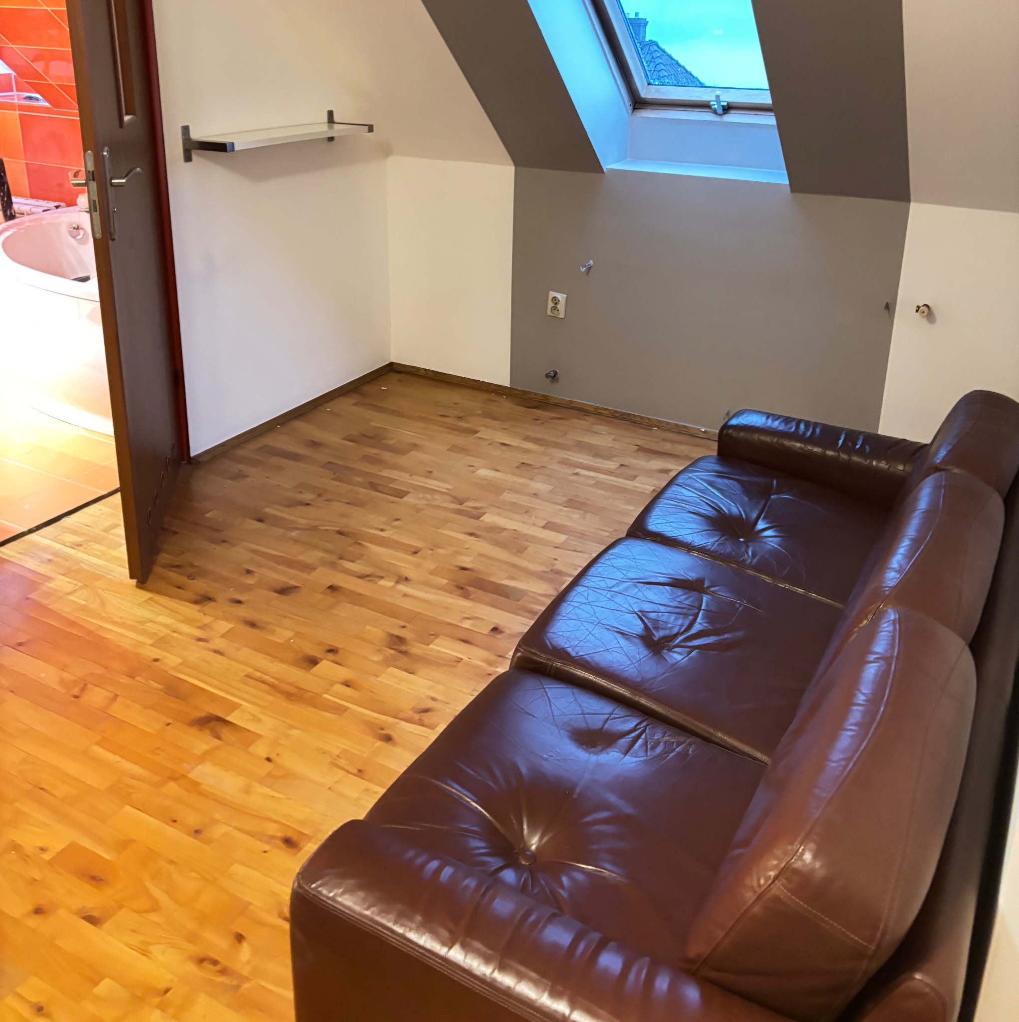 Poddasze po remoncie: parkiet z jasnego drewna, szara ściana pod oknem dachowym, brązowa skórzana sofa. Widok na łazienkę z wanną.