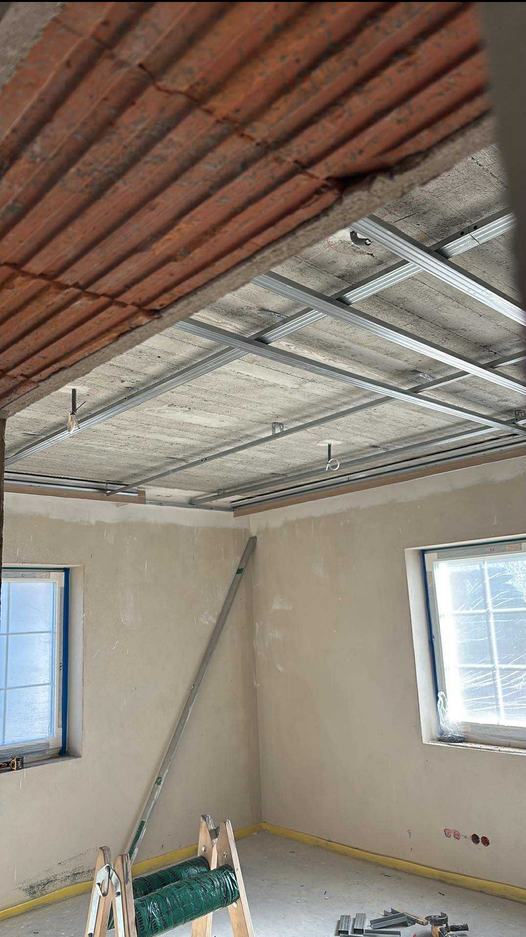 Montaż stelaża sufitu podwieszanego z płyt karton-gips w pomieszczeniu z oknami zabezpieczonymi folią. Widoczna drabina i narzędzia.