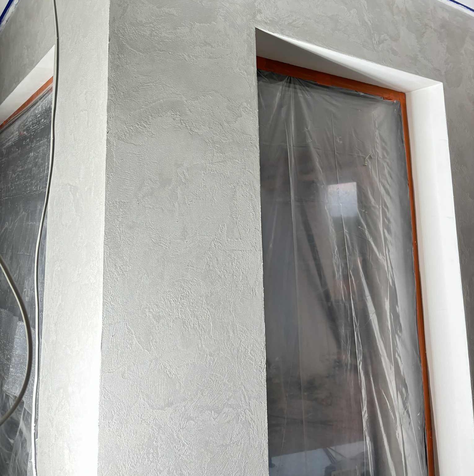 Ściana z dekoracyjną, szarą strukturą tynku, zabezpieczone okno folią malarską. Detal wykończenia wnętrza z bliska, ukazujący fakturę i przygotowanie do dalszych prac.