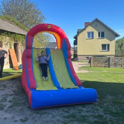 Dmuchana Zabawa - Dziewczynka z radością zjeżdża na dmuchanej zjeżdżalni w kolorach czerwieni, żółci i błękitu. W tle dom i zieleń trawnika w słoneczny dzień.