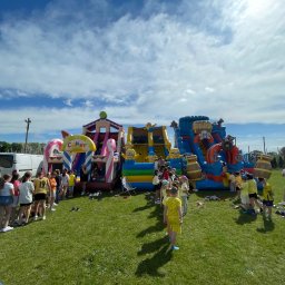 Dmuchana Zabawa - Kolorowe dmuchańce z motywami cukierków, Minionków i piratów ustawione na trawie, otoczone grupą dzieci czekających w kolejce. W tle biały van i wóz strażacki.