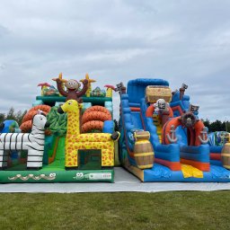 Dmuchana Zabawa - Dwa dmuchańce: dżungla ze zwierzętami (zebra, żyrafa, małpa) i piracki statek ze zjeżdżalnią, ustawione na trawie. W tle biały van.