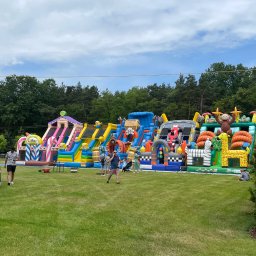 Dmuchana Zabawa - Kolorowy park dmuchany z motywami zwierząt i postaciami z bajek, ustawiony na trawniku podczas imprezy plenerowej. Widoczni ludzie korzystający z atrakcji.