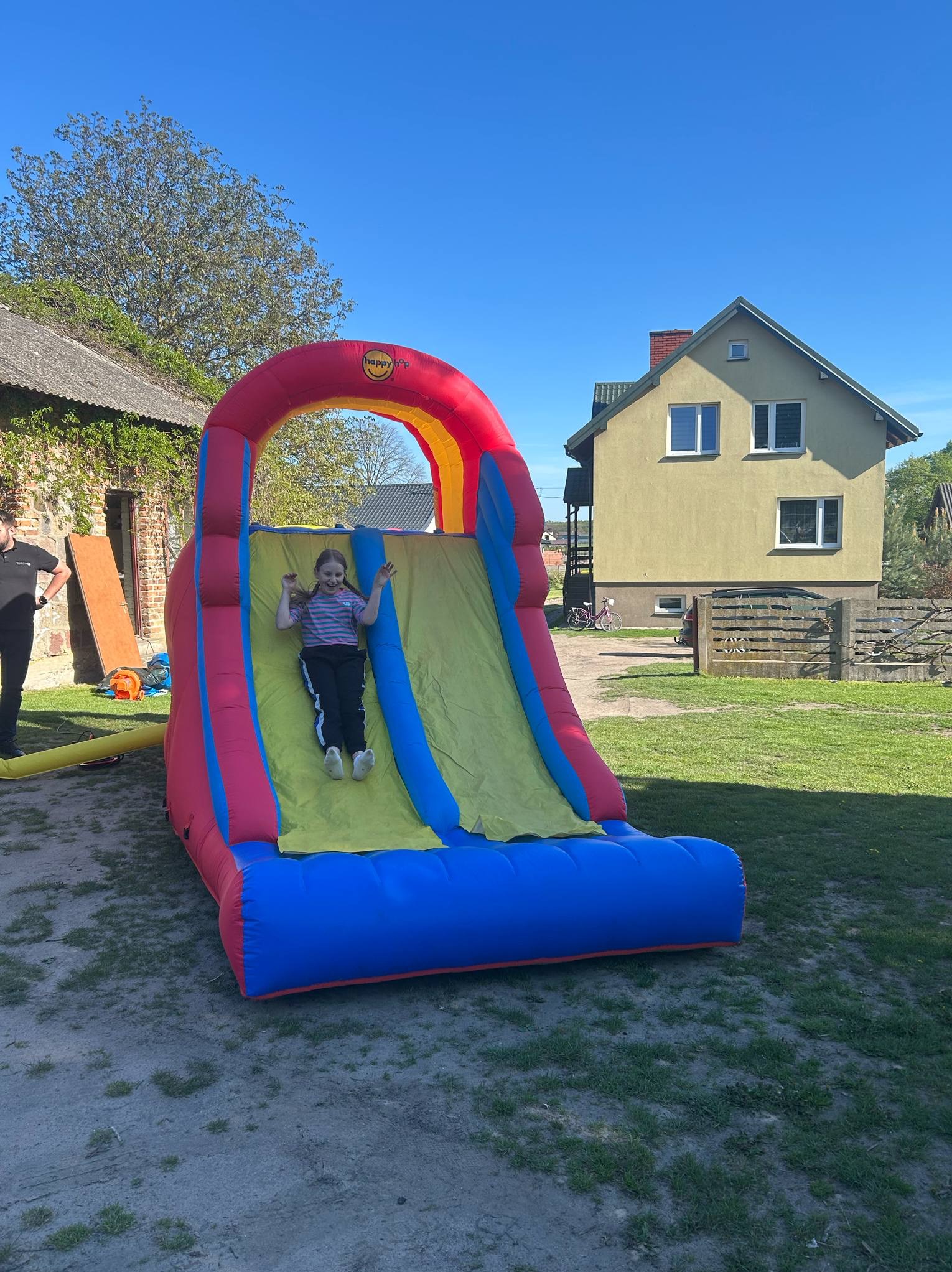 Dziewczynka z radością zjeżdża na dmuchanej zjeżdżalni w kolorach czerwieni, żółci i błękitu. W tle dom i zieleń trawnika w słoneczny dzień.