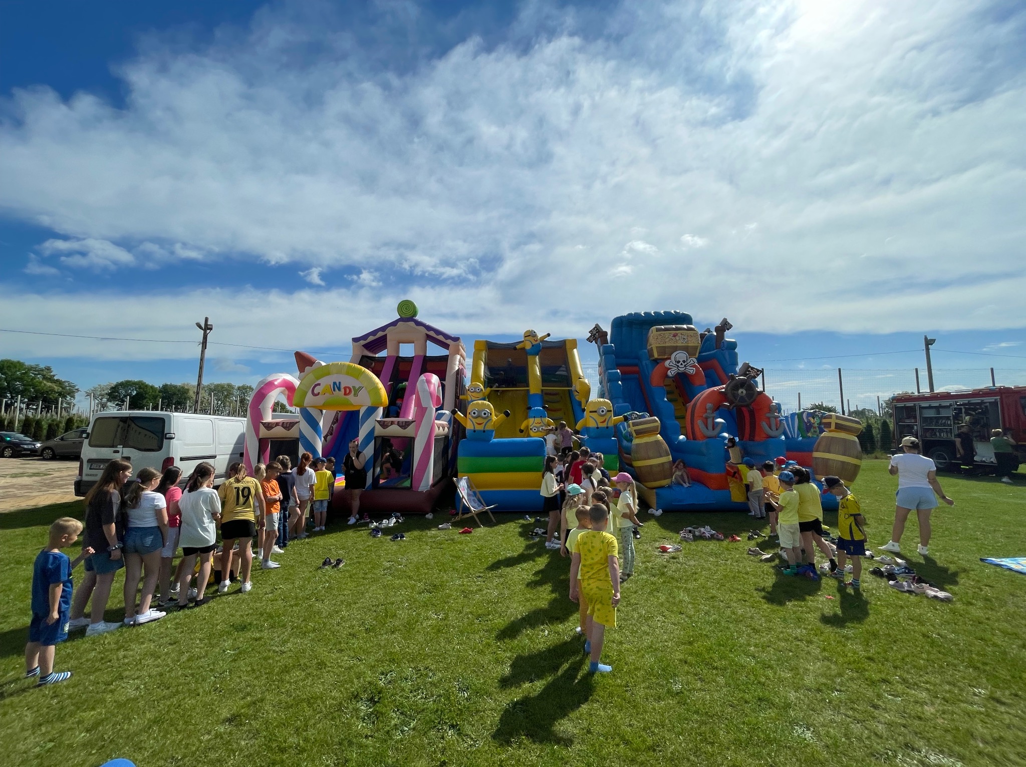 Kolorowe dmuchańce z motywami cukierków, Minionków i piratów ustawione na trawie, otoczone grupą dzieci czekających w kolejce. W tle biały van i wóz strażacki.