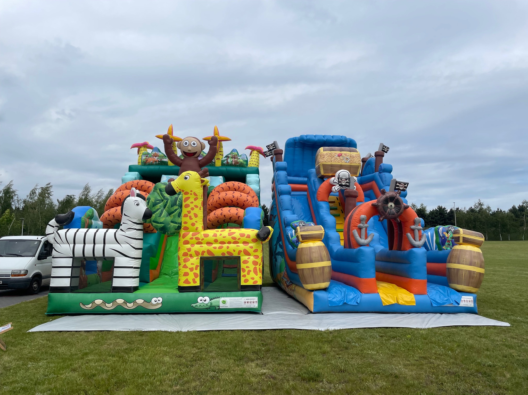 Dwa dmuchańce: dżungla ze zwierzętami (zebra, żyrafa, małpa) i piracki statek ze zjeżdżalnią, ustawione na trawie. W tle biały van.