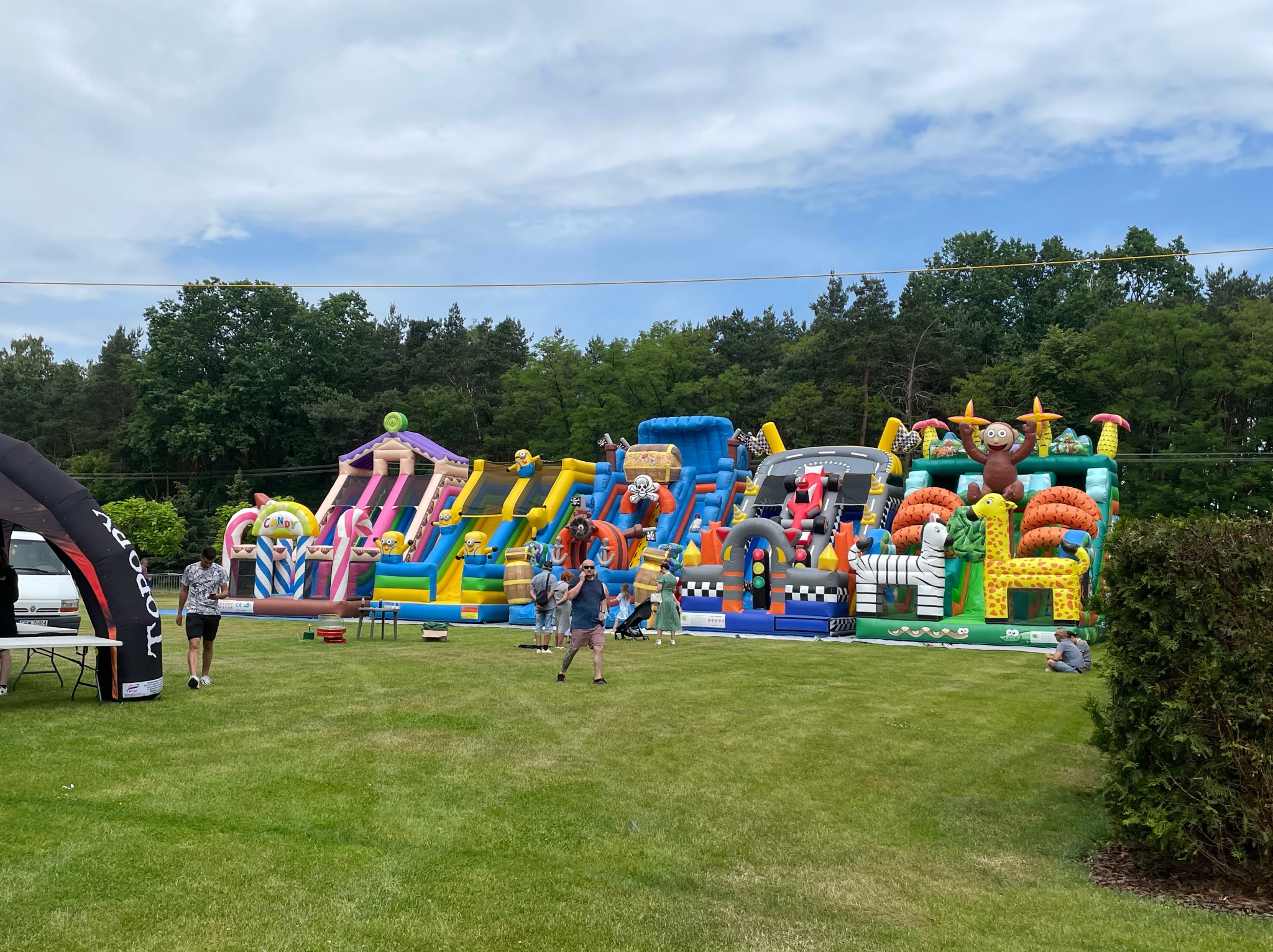 Kolorowy park dmuchany z motywami zwierząt i postaciami z bajek, ustawiony na trawniku podczas imprezy plenerowej. Widoczni ludzie korzystający z atrakcji.