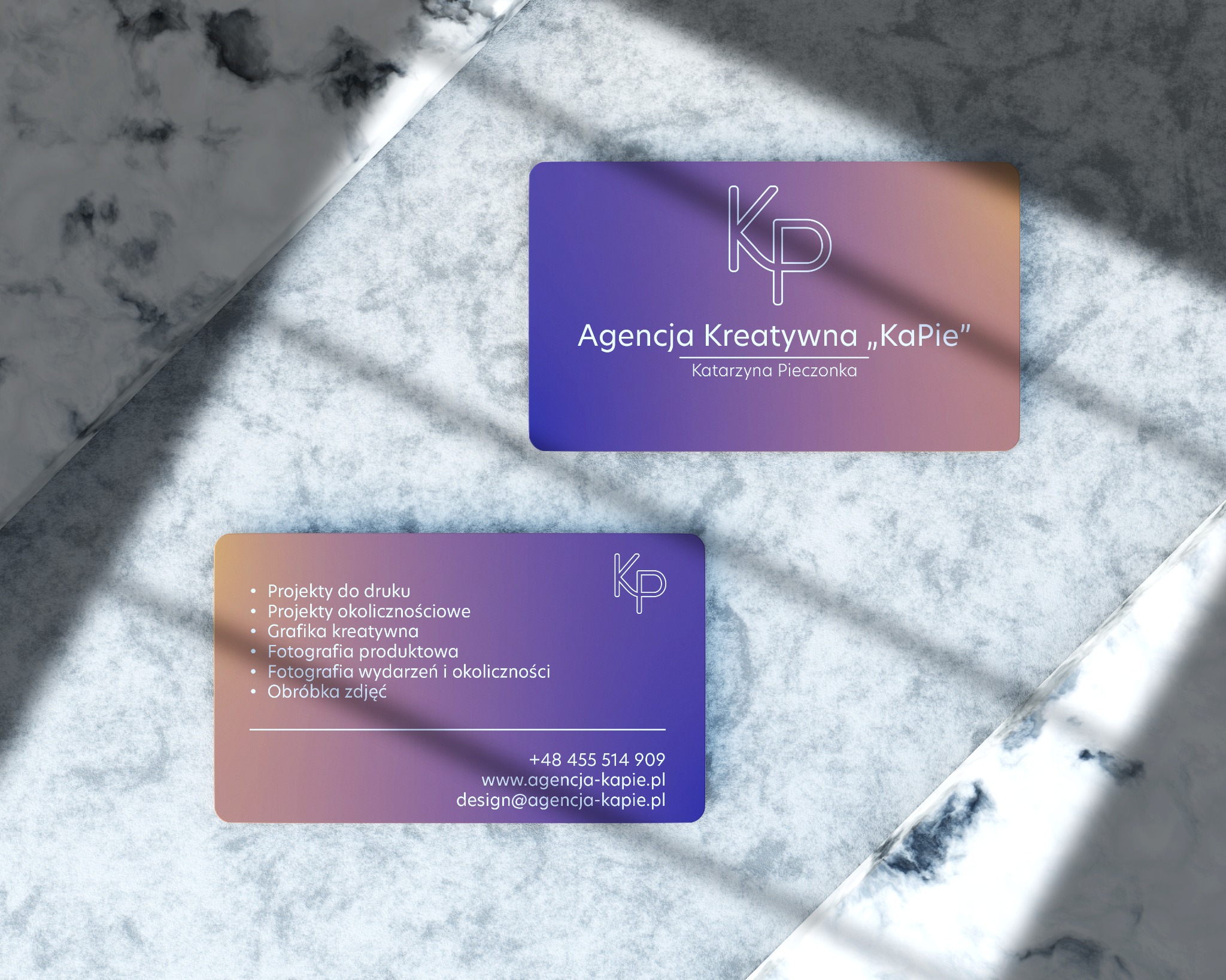 Dwie wizytówki Agencji Kreatywnej „KaPie” na marmurowym tle w cieniu. Gradient fioletowo-pomarańczowy, logo KP, dane kontaktowe i lista usług.
