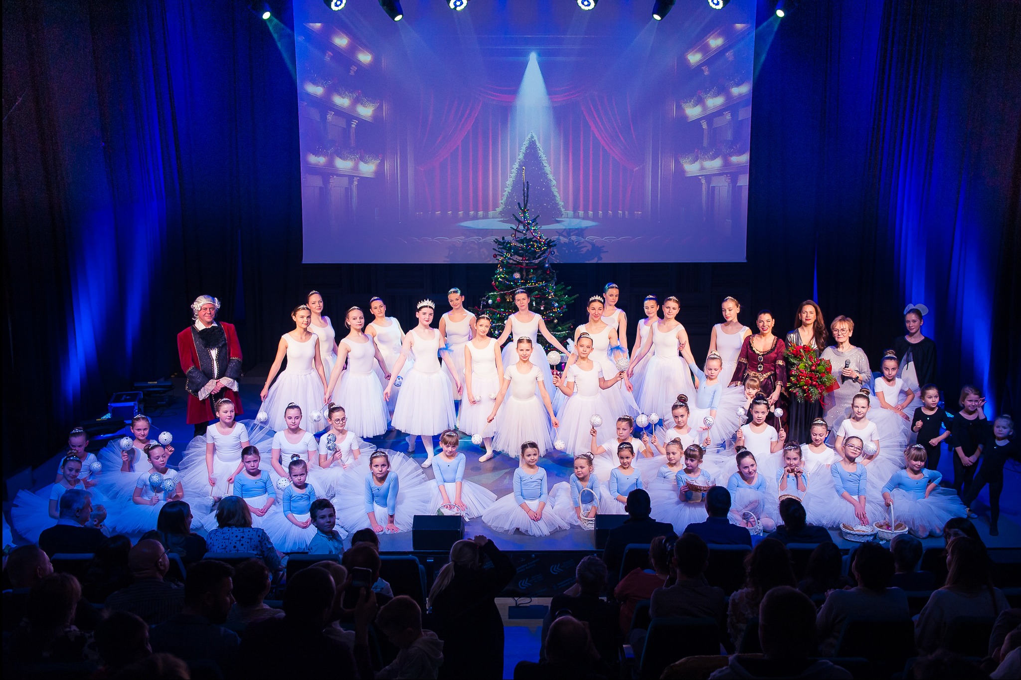 Grupowe zdjęcie baletnic na scenie, w tle dekoracja świąteczna i publiczność. Widoczny mężczyzna w kostiumie z epoki i kobiety z bukietem kwiatów. Scena oświetlona reflektorami.