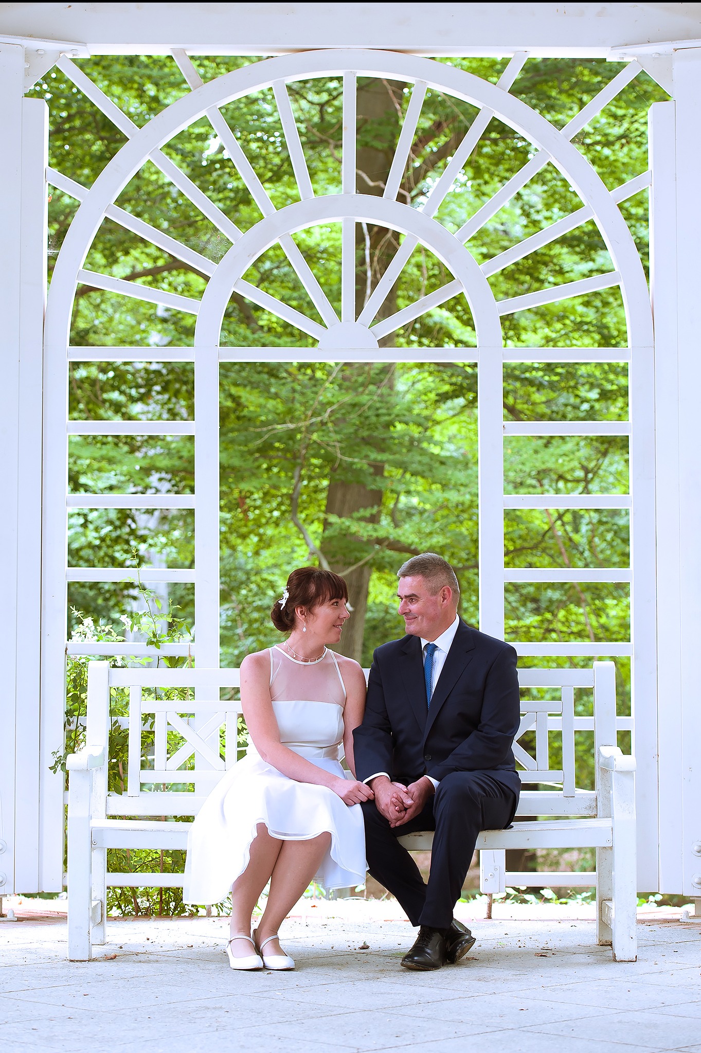 Para młoda siedzi na białej ławce w altanie z ażurową konstrukcją, spoglądając na siebie z uśmiechem. Romantyczna sesja plenerowa w otoczeniu zieleni.