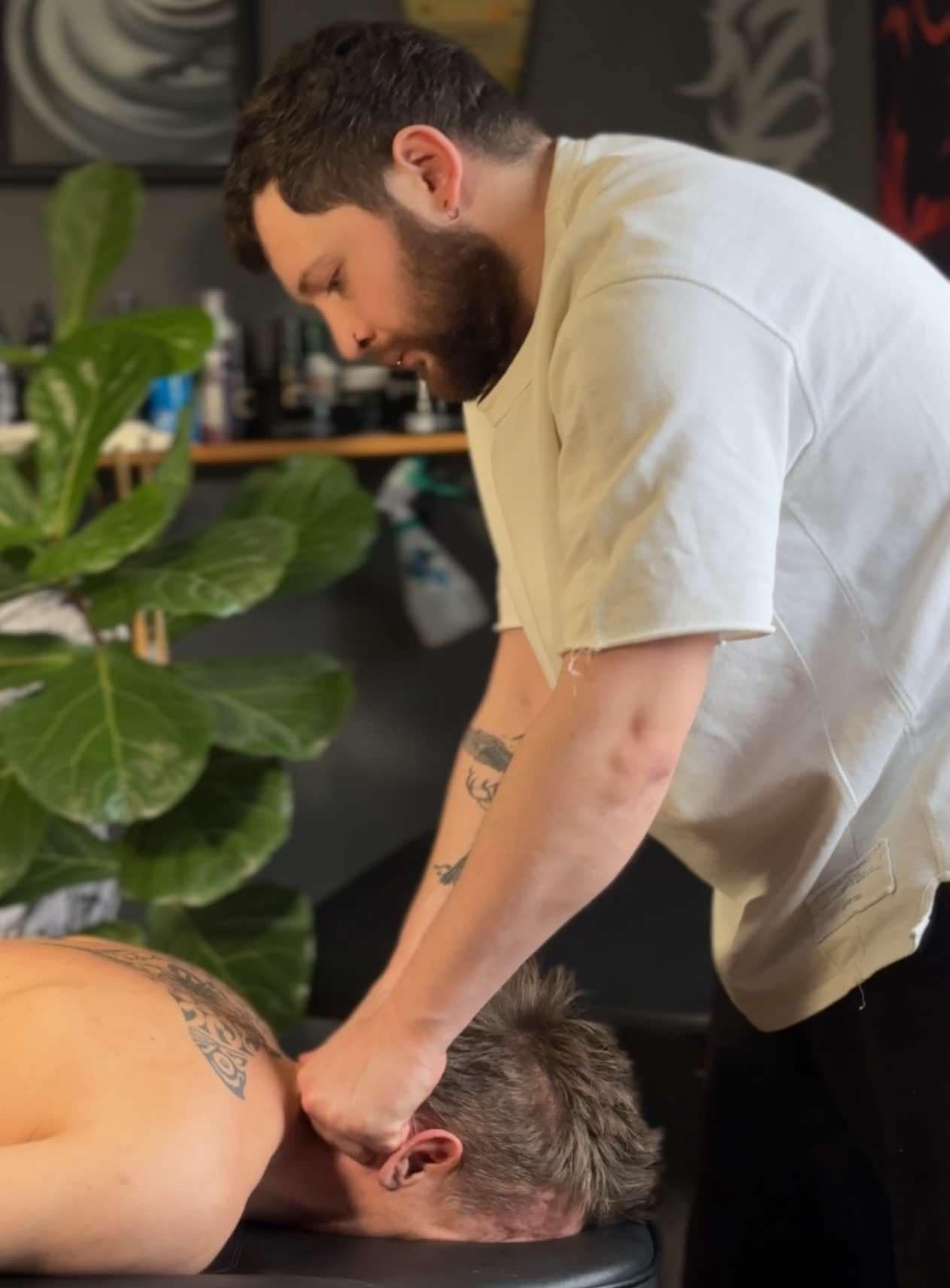 Mężczyzna z brodą w białej koszulce wykonuje masaż karku klientowi leżącemu na czarnym stole do masażu, w tle widoczna roślina doniczkowa i półka.