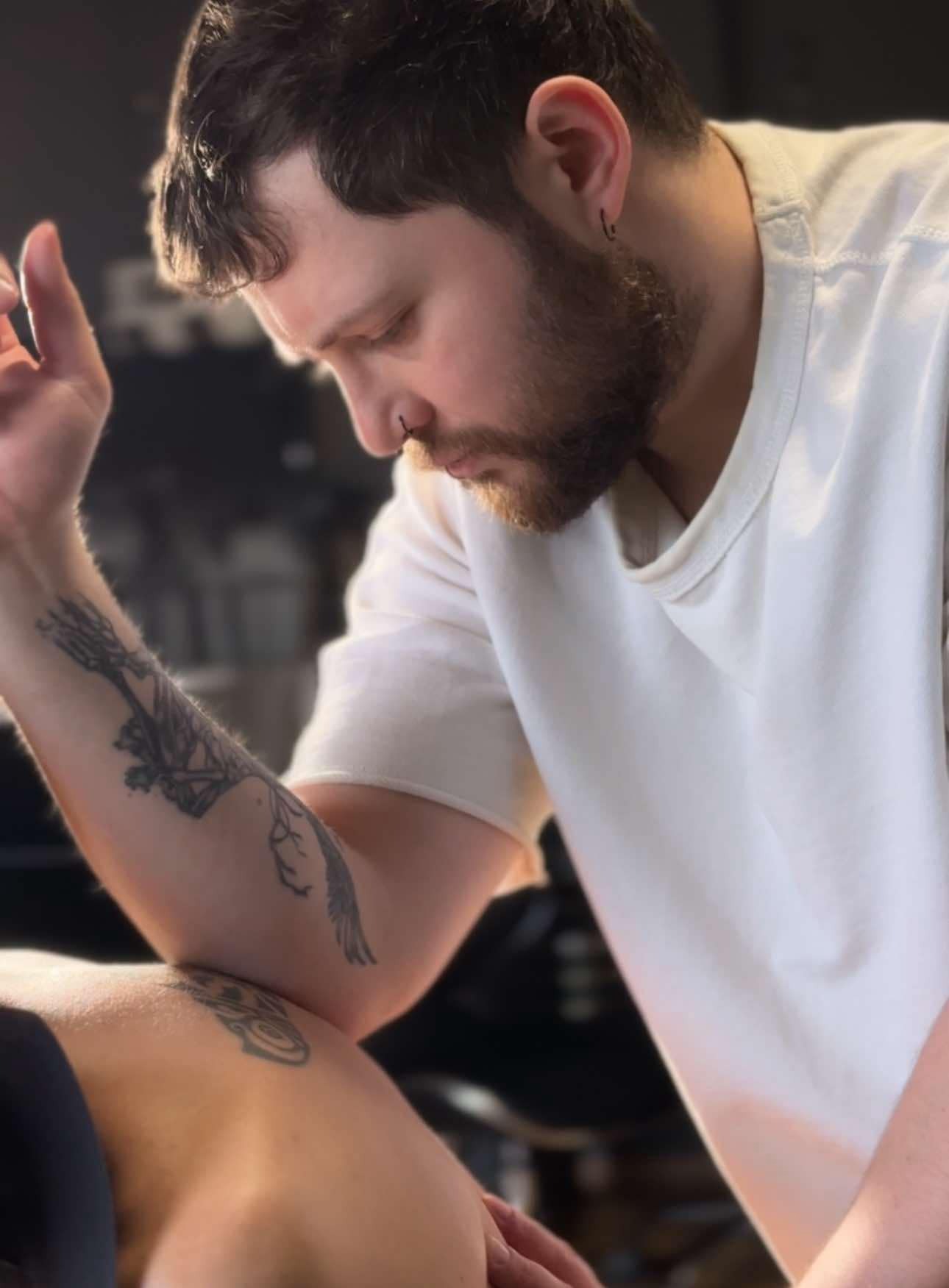 Mężczyzna z tatuażami na ramieniu wykonuje masaż pleców, pochylony nad klientem. Widoczny fragment pleców klienta i dłoń masażysty.