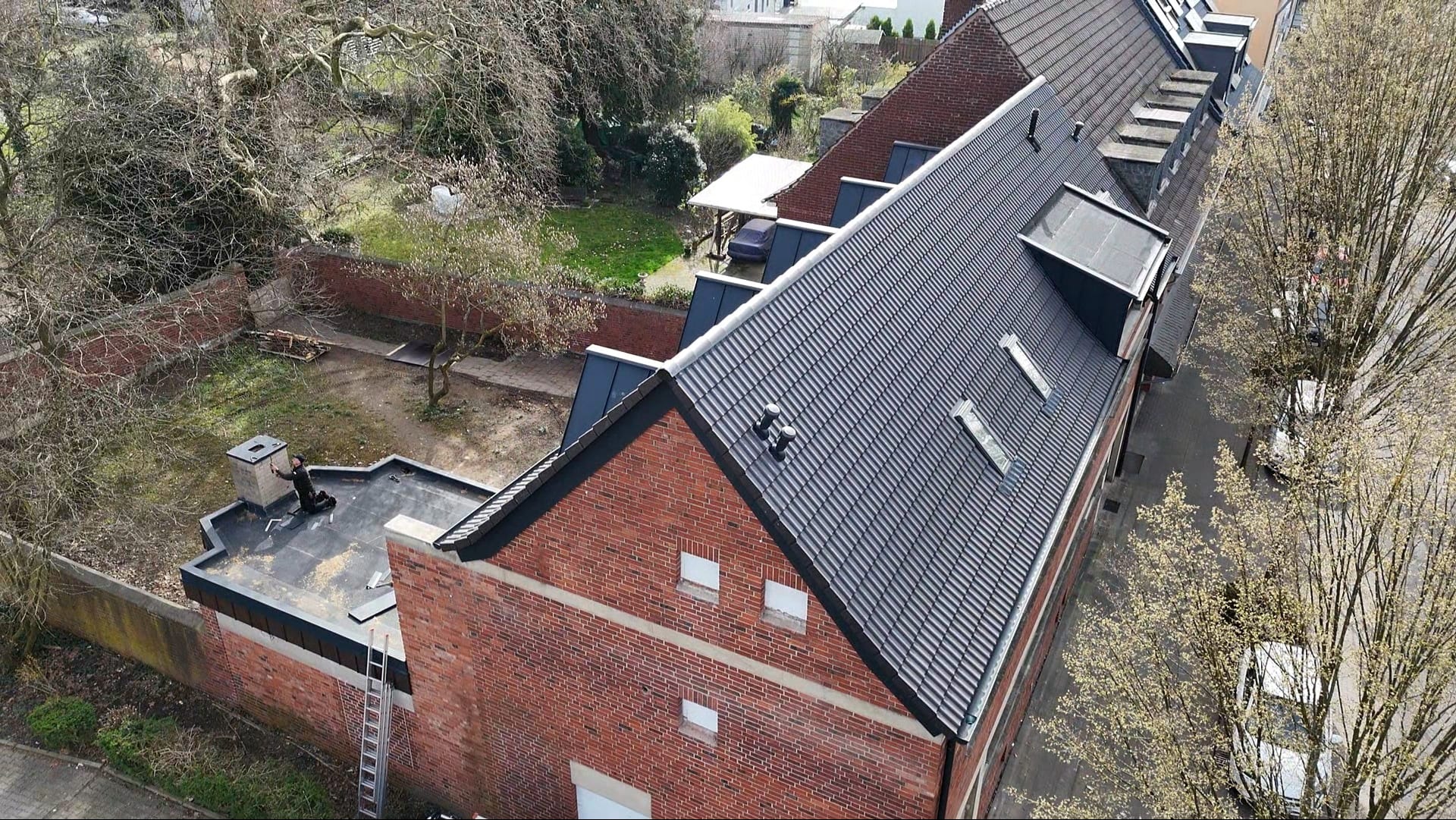 Widok z góry na budynek z ciemną dachówką, ceglaną elewacją i płaskim dachem z kominem, na którym stoi osoba. W tle ogród i ulica z drzewami.