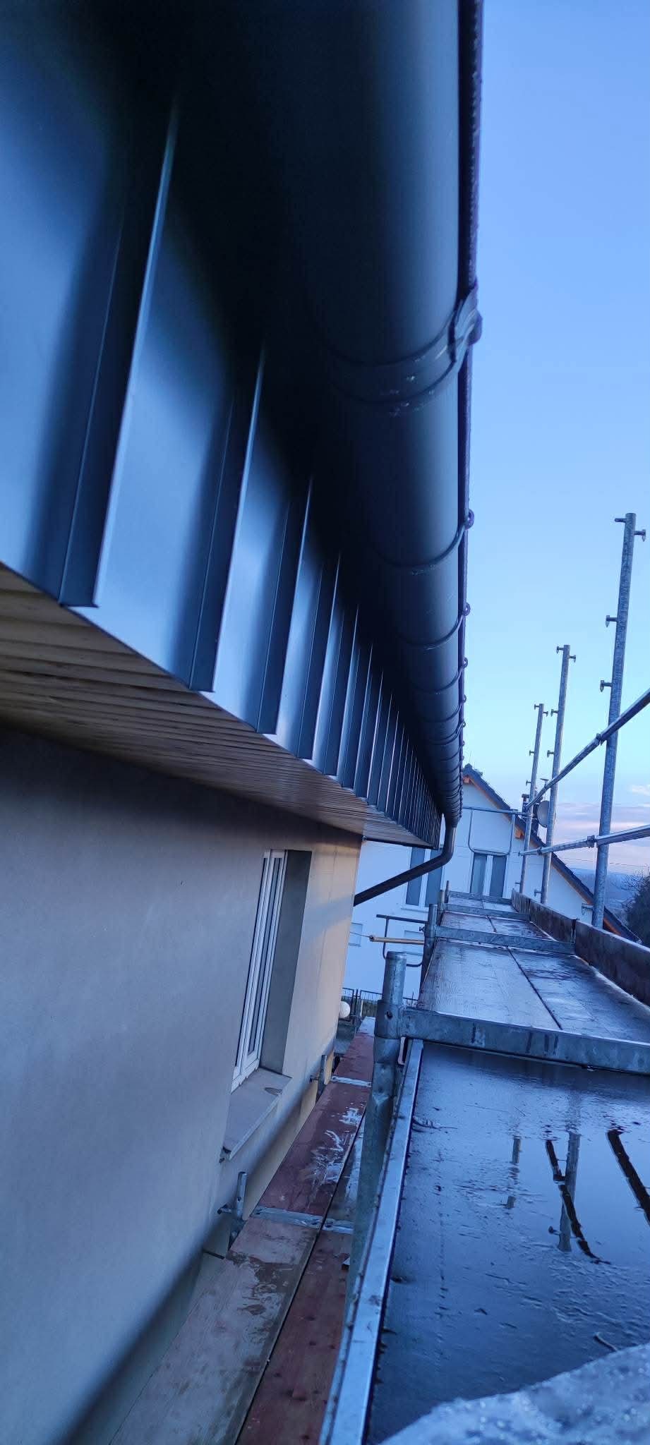 Zamontowana, ciemna rynna dachowa z blachy, widoczna z rusztowania. Ujęcie z dołu, na tle błękitnego nieba. Widoczne elementy mocujące rynnę.