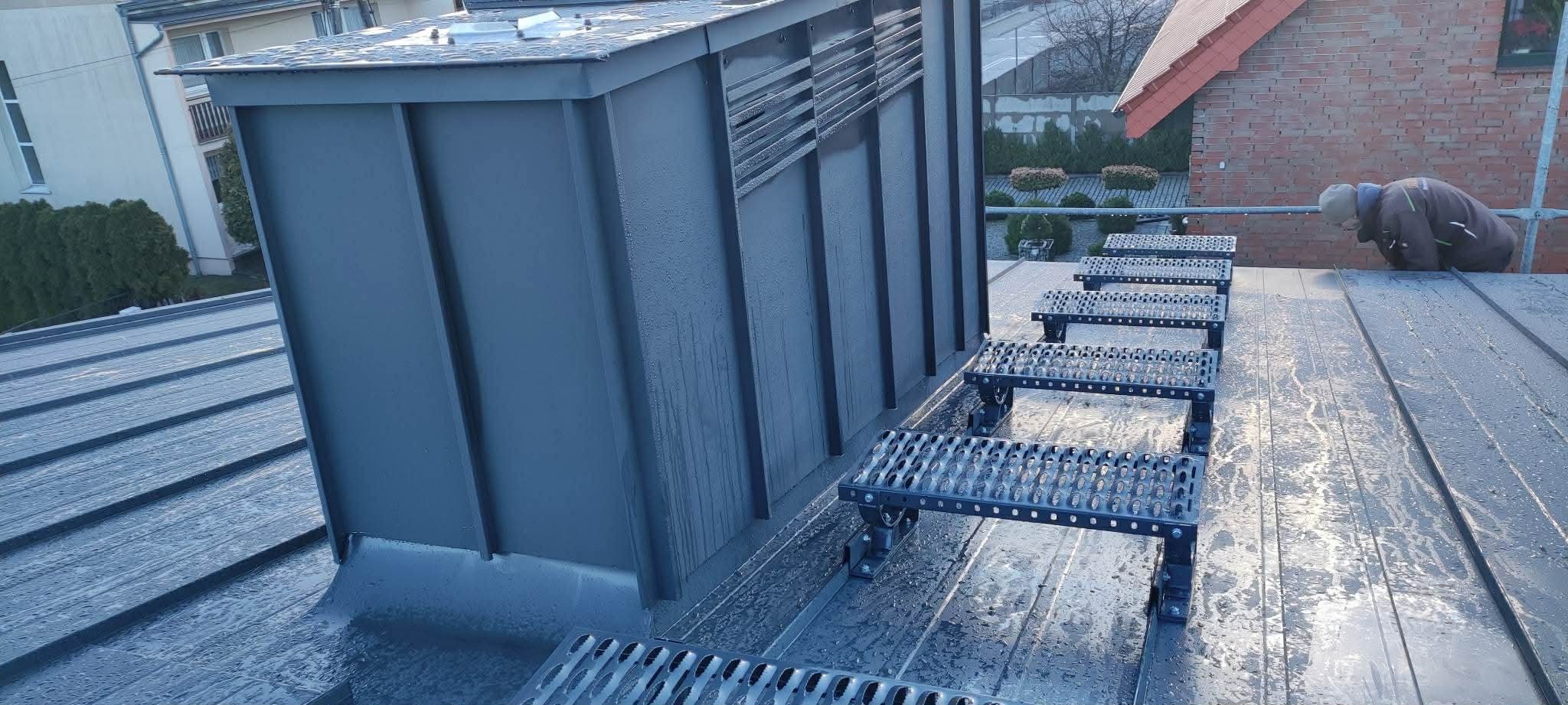 Szary dach z blachy trapezowej z zamontowanymi schodami dachowymi i kominem wentylacyjnym. Widoczny dekarz w oddali.