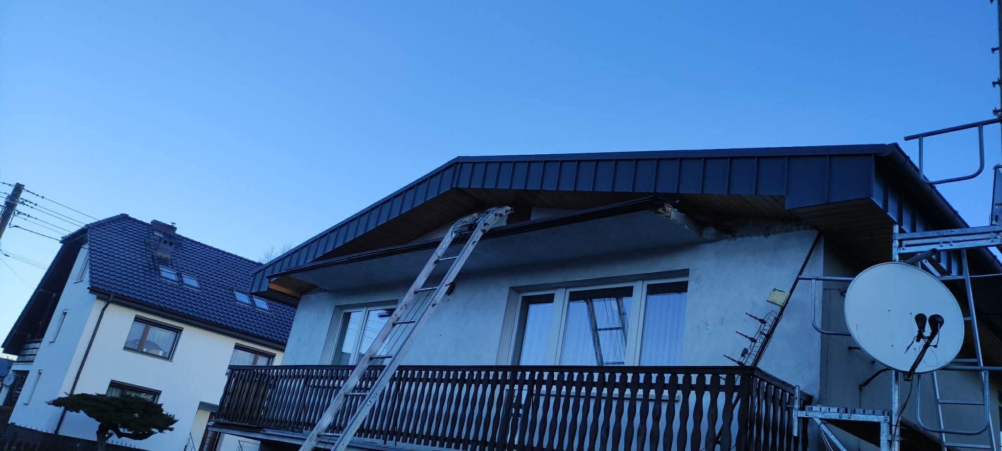Dom z balkonem i nowym, ciemnym dachem. Przystawiona drabina sugeruje prace dekarskie. Widoczna antena satelitarna i fragment drugiego budynku w tle.