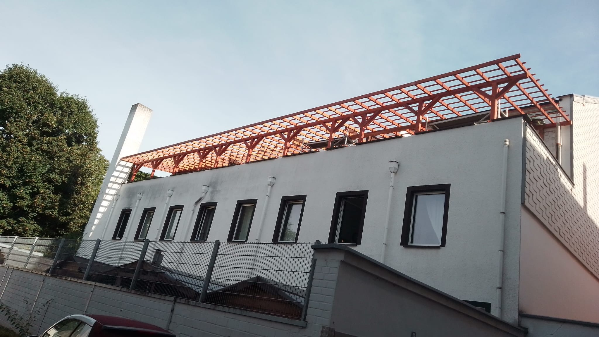 Biały budynek z ciemnymi oknami i drewnianą konstrukcją na dachu, widziany z dołu pod kątem. Widoczne rynny i fragment ogrodzenia.