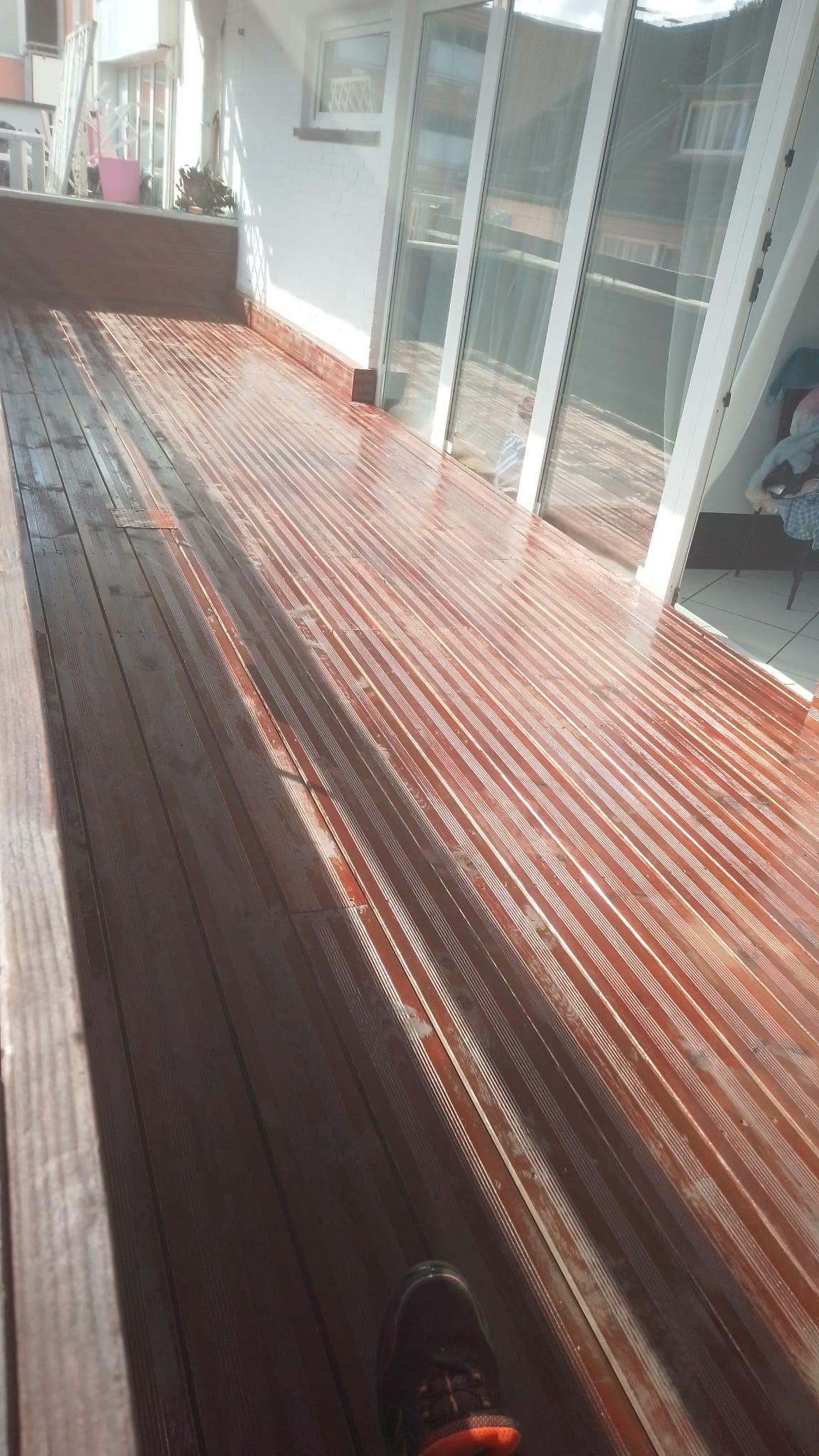 Błyszczący, drewniany taras po impregnacji, z widocznymi refleksami światła i fragmentem buta w rogu. Widok na przeszklone drzwi balkonowe i białą ścianę.