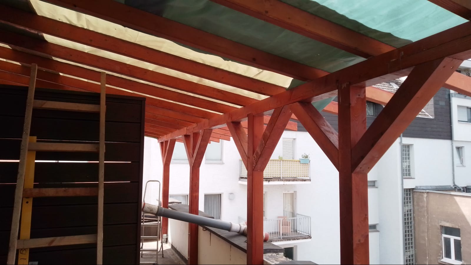 Drewniana konstrukcja zadaszenia balkonu w trakcie budowy, widok od dołu, z elementami zabezpieczenia i drabiny. W tle budynki mieszkalne.