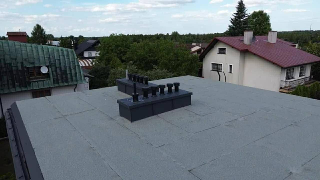Szary, płaski dach pokryty papą termozgrzewalną z kominami wentylacyjnymi na tle zabudowy jednorodzinnej i zieleni. Widok z góry, w słoneczny dzień.