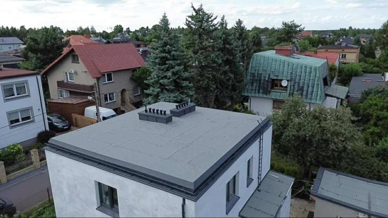 Szary, płaski dach budynku z kominami wentylacyjnymi na tle zabudowy jednorodzinnej i zieleni. Widok z góry.
