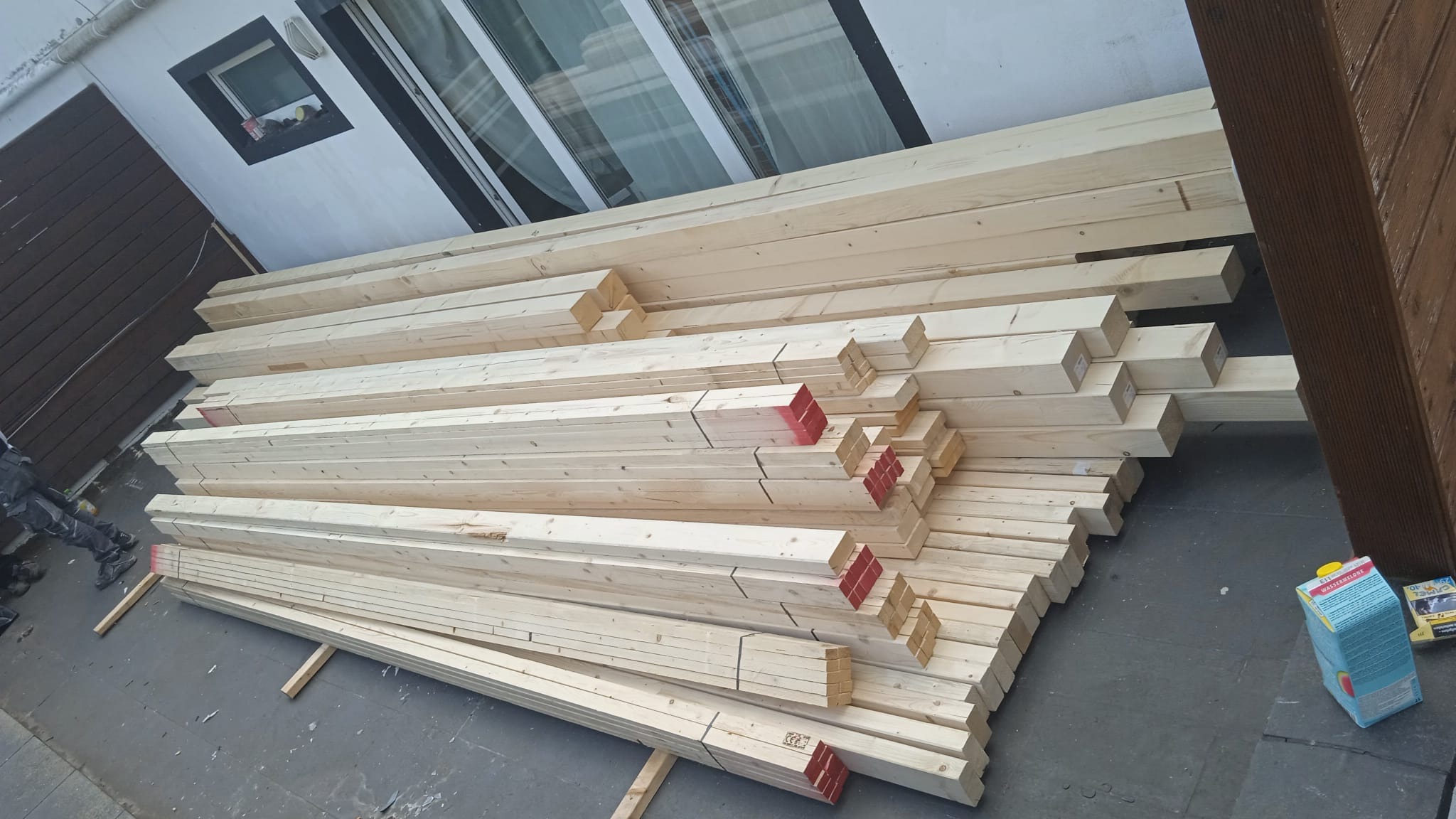 Stos belek drewnianych z czerwonymi oznaczeniami na tle ściany budynku z oknem i fragmentem brązowej elewacji. Przy belkach karton soku i opakowanie po paście.