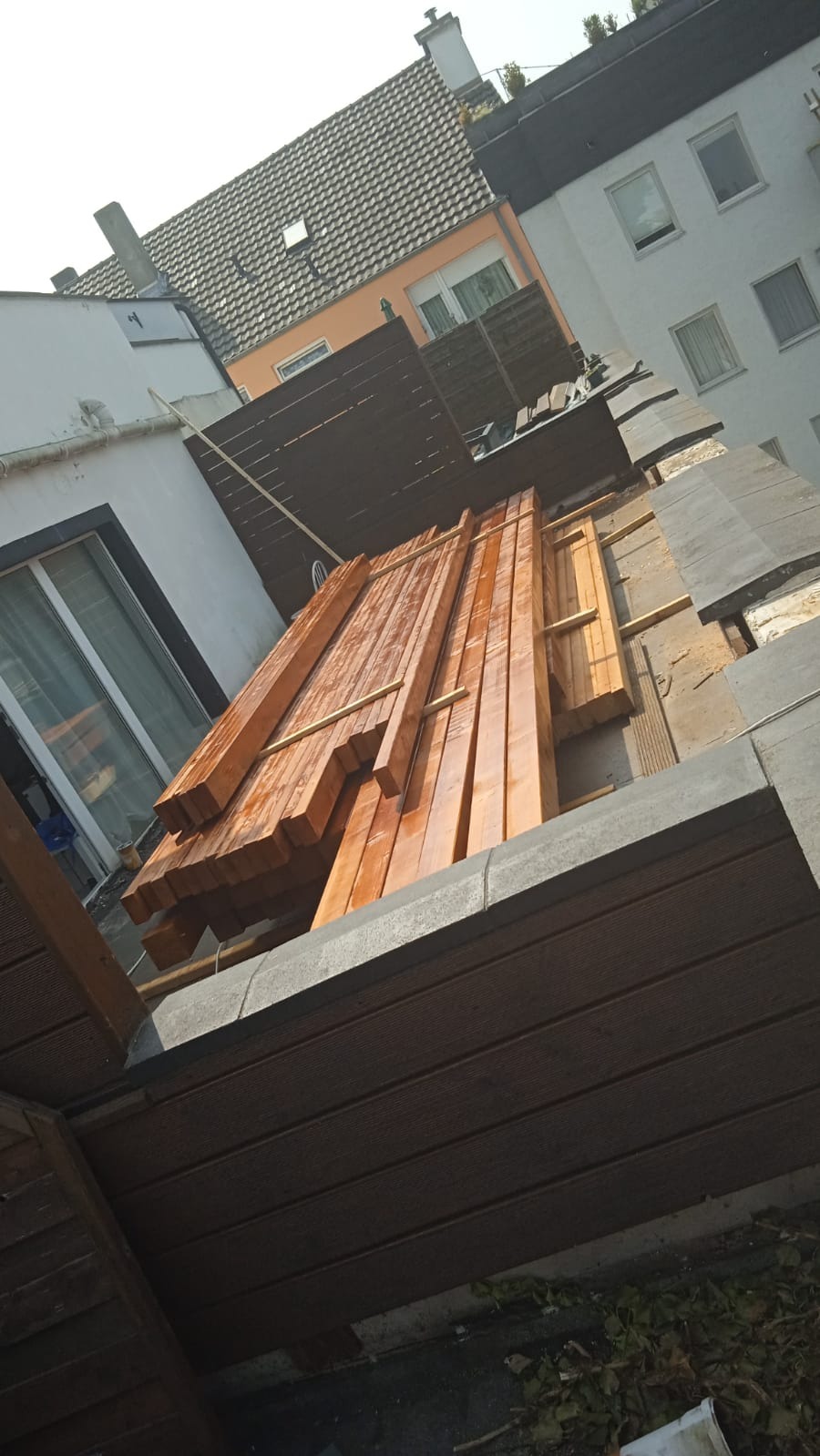 Przygotowane, impregnowane drewno na dachu budynku wielorodzinnego, widoczne elementy konstrukcyjne i fragmenty zabudowy. Ujęcie z dołu, w tle budynki mieszkalne.